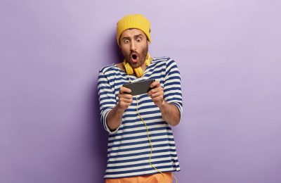 Photo of shocked unshaven guy holds mobile phone horizontally, holds breath, plays games online, being addicted, wears yellow hat and striped jumper, stands in studio against purple background